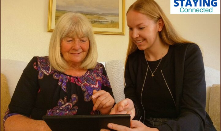 Hannah, sitting on a sofa beside Mary, an 86-year-old woman. Hannah is gently showing Mary how to use a tablet.