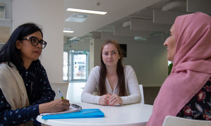 Three women are sitting at a table, engaged in conversation. One woman on the left is wearing glasses and taking notes.